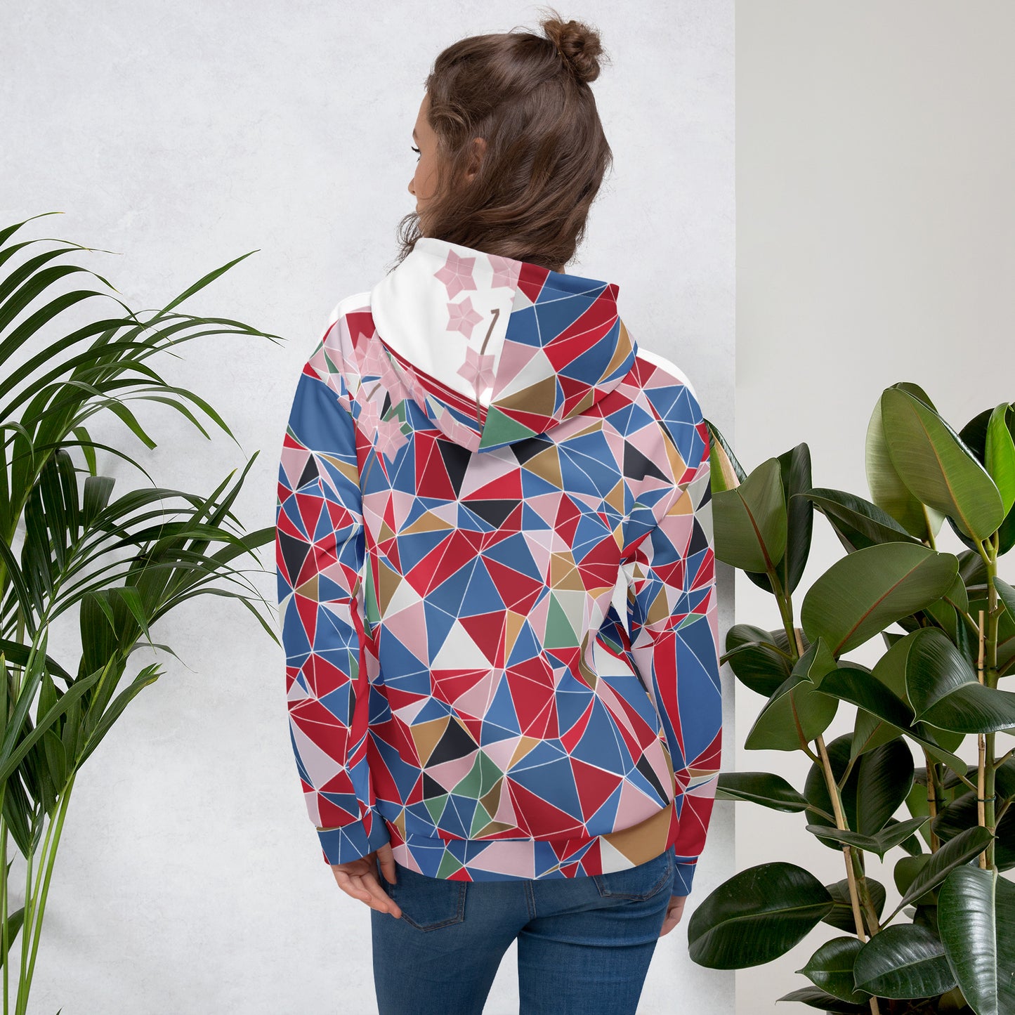 Person wearing a colorful geometric-patterned hoodie standing between two plants against a white wall.