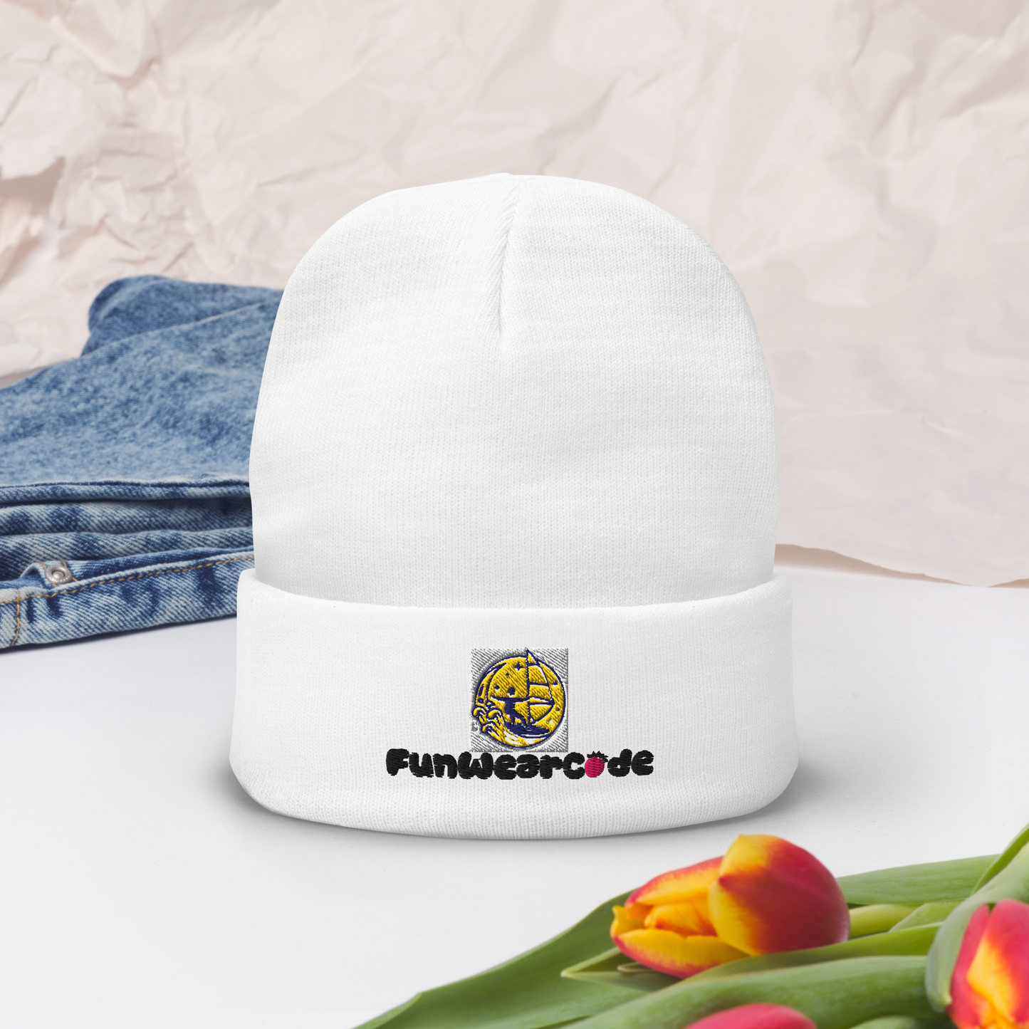 White beanie with 'FunWearCode' logo and text on a white surface with tulips and denim jeans.